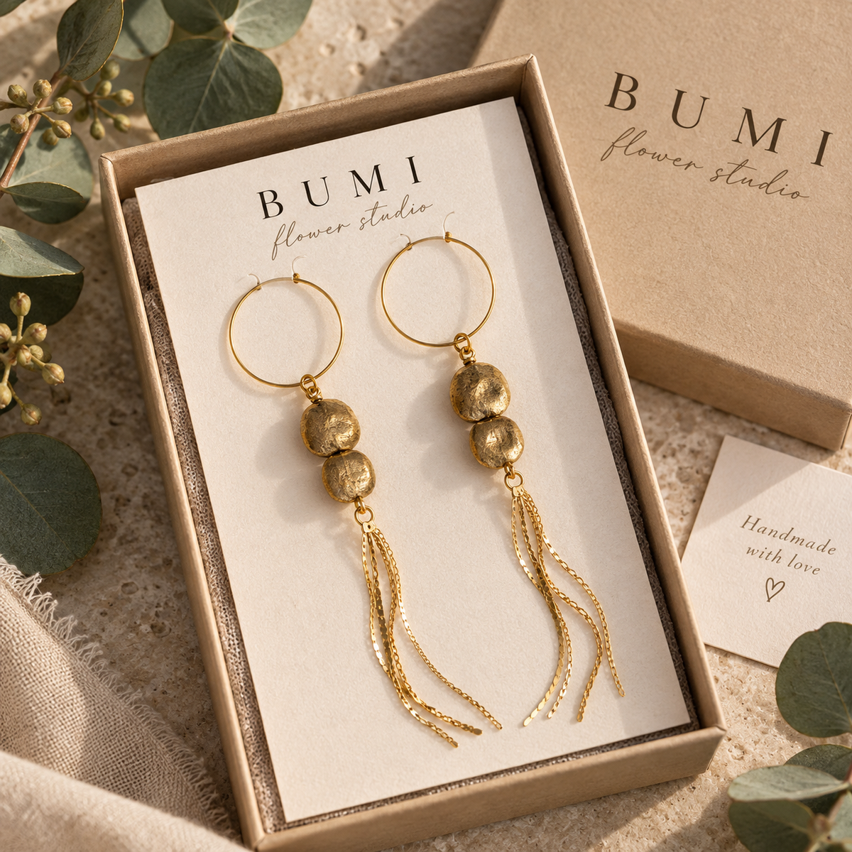 Gold-toned dangle gumnut earrings displayed in a beige gift box with a cream insert card printed with the brand name BUMI flower studio. The earrings have circular hoops, real gumnuts, and long chain tassels.