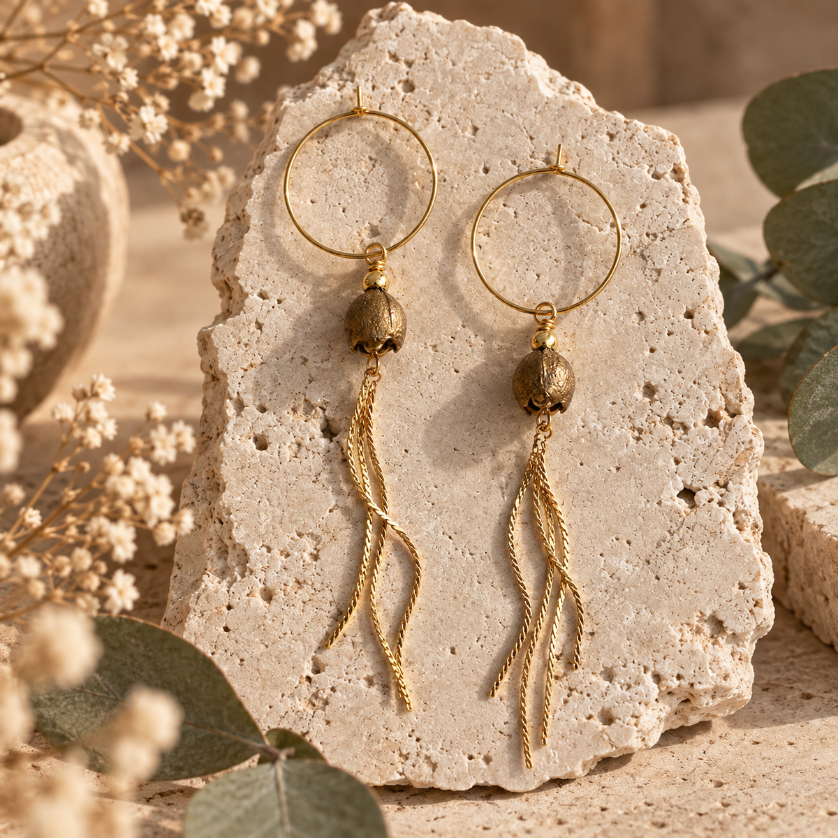 A pair of gold-toned gumnut dangle earrings displayed on a textured stone slab, surrounded by dried flowers and green leaves in a warm, natural setting.
